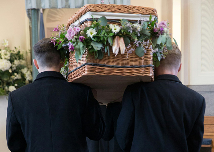 Two men in black suits carrying a flower-adorned wicker coffin symbolizing parents disowning their kids.