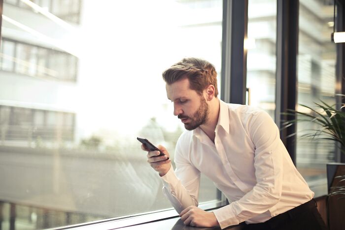 Man in white shirt sitting by window, focused on phone screen, representing red flag poll concept.