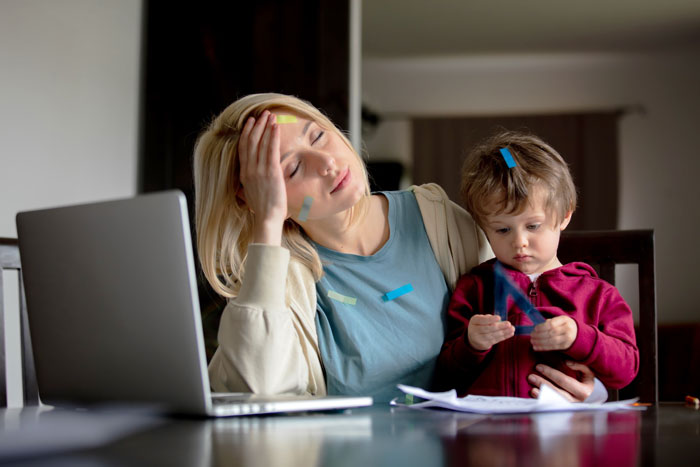 Tired woman multitasking babysitting neighbor's kid with colorful sticky notes on her face inside a home.