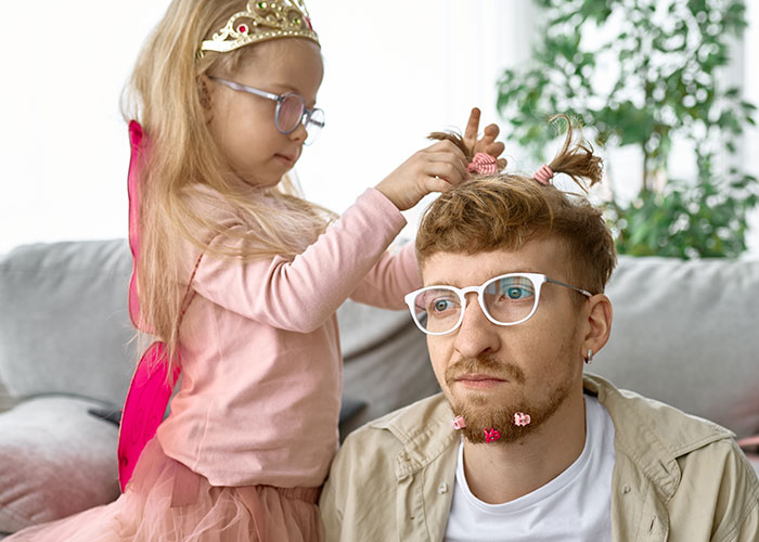 Man with glasses looking tired while a little girl plays hairstylist in a cozy living room setting.