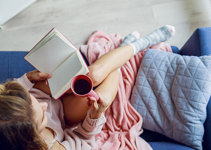Person relaxing on couch with coffee and a book, illustrating man refusing to babysit roommate&rsquo;s kid after night shift.