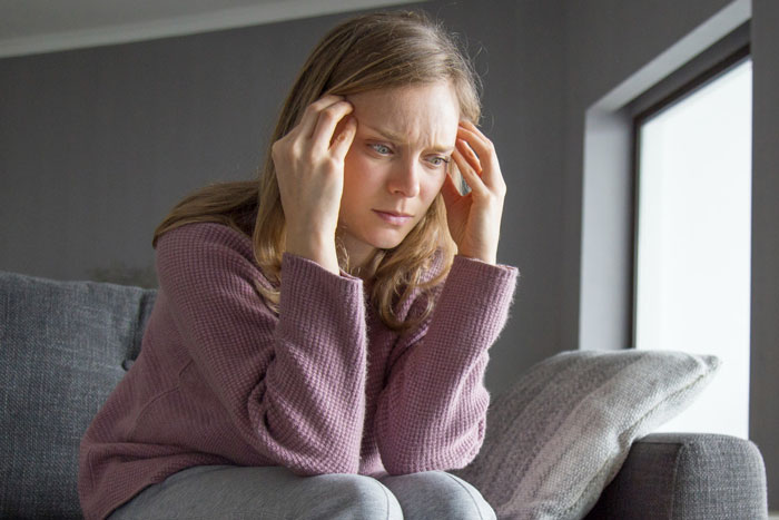 Woman sitting on couch, looking stressed and worried while holding her head near a window in a dimly lit room.