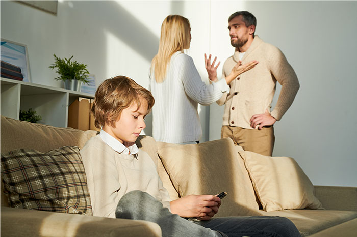 Child sitting on couch looking at phone while parents in background argue about kids&rsquo; diet and custody arrangements.