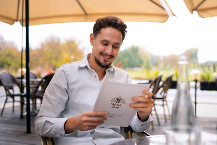 Man smiling while reading a menu at an outdoor restaurant, relating to friend laughs off vegan warning story.