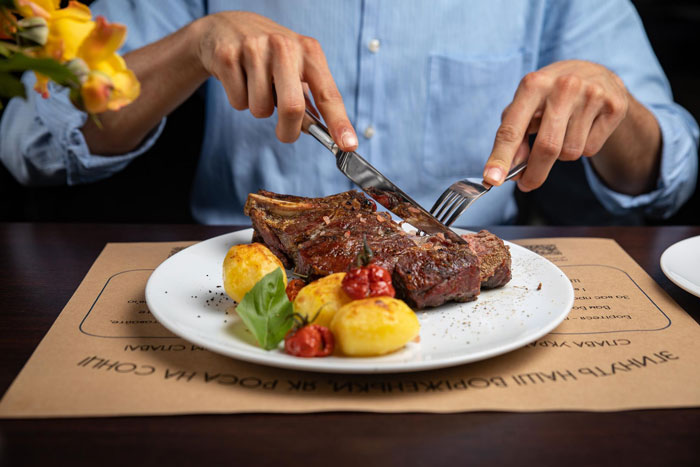 Person cutting steak with fork and knife, highlighting friend laughs off vegan&rsquo;s warning about paying for his food.