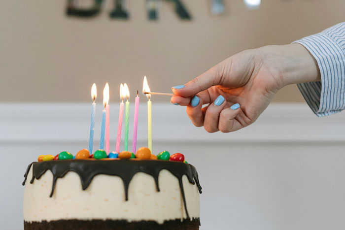 Hand with blue nail polish lighting candles on birthday cake with colorful candies and chocolate drip frosting. Hand with blue nail polish lighting candles on birthday cake with colorful candies and chocolate drip frosting.