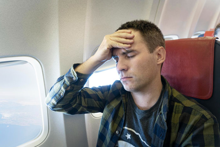 Man on plane with hand on forehead, appearing stressed while reflecting on life-changing trip after cancer battle.