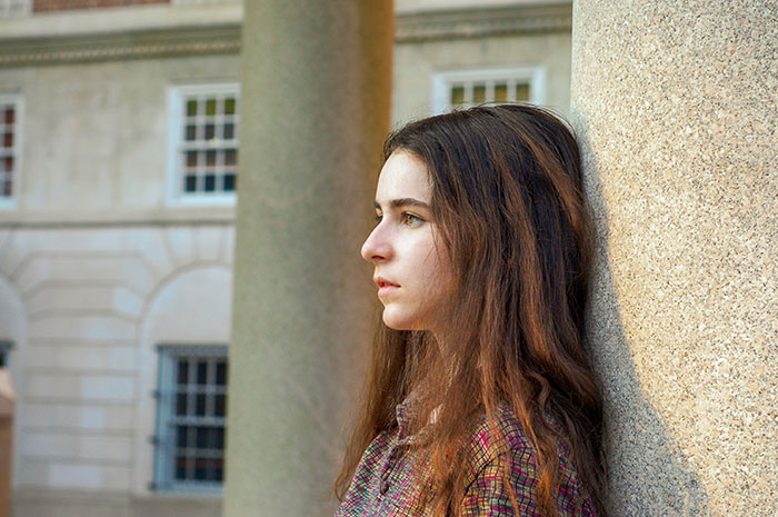Young woman leaning on a stone column outside, reflecting on removing phone tracking used by her parents to see location.