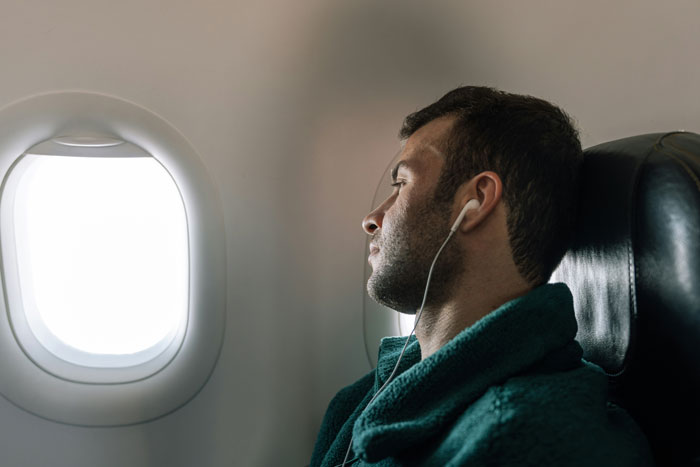 Passenger moves seats on flight, wearing earphones, looking out airplane window, affected by nearby special needs kid's kickfest.