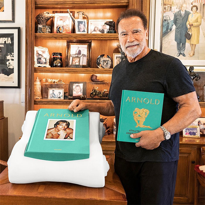 Actor Arnold Schwarzenegger holding a green book, surrounded by memorabilia, representing richest actors in the world 2025.