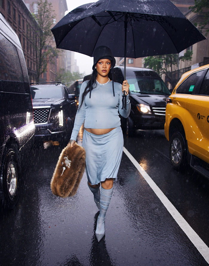 Rihanna shows her 3rd pregnancy in a blue outfit holding an umbrella, walking on a rainy city street.