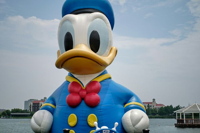 Giant inflatable Donald Duck wearing a blue sailor suit with red bowtie near waterfront under cloudy sky.