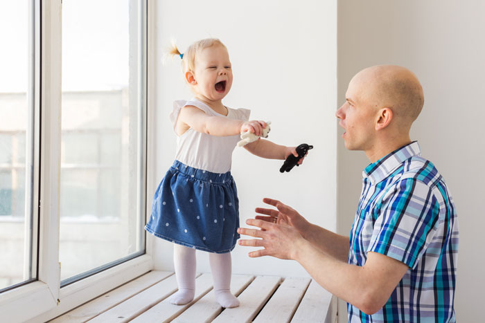 Man interacts with toddler daughter at a window, capturing the challenge of a roommate vanishing with ex and leaving a kid behind.