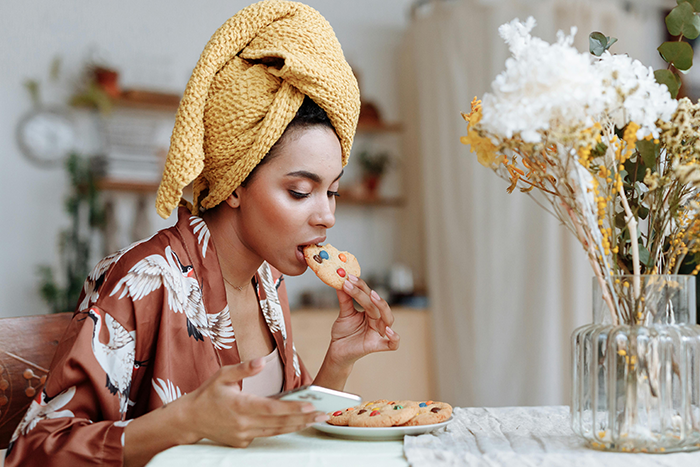 Middle-aged woman with a shopping habit eating cookies, holding a phone, in a cozy home setting with flowers on the table.