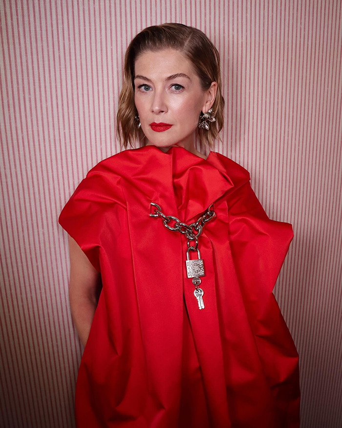 Rosamund Pike in a red dress with a padlock chain, posing against a striped background related to Bond girl auditions.