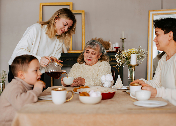 Young woman serving tea to an elderly woman while two others sit at a table, reflecting family and kids dynamics conversation.