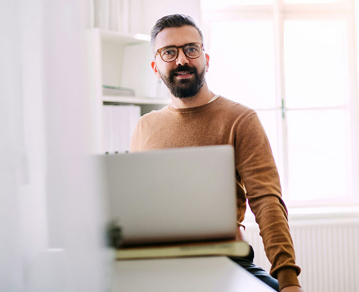 Man with glasses and beard sitting at desk with laptop, portraying confidence in a tall and loud office setting.