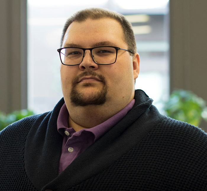 Man wearing glasses and a purple shirt with a dark sweater, confidently facing the camera with tall and loud demeanor.
