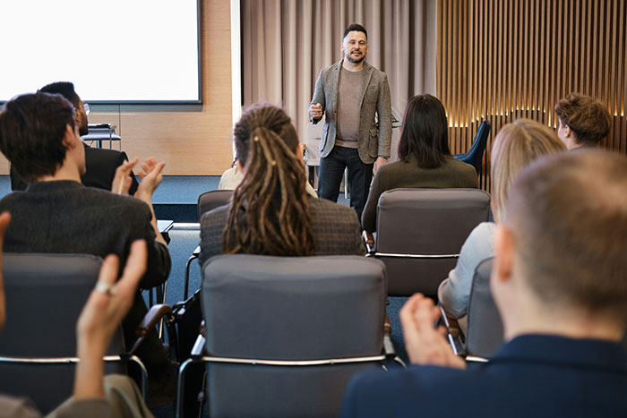 Man standing and speaking confidently to an applauding audience about comebacks to entitled bosses in a modern office setting