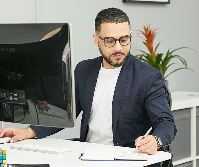 Man wearing glasses and a blazer working at a desk with computer and notebook, portraying confident boss attitude.