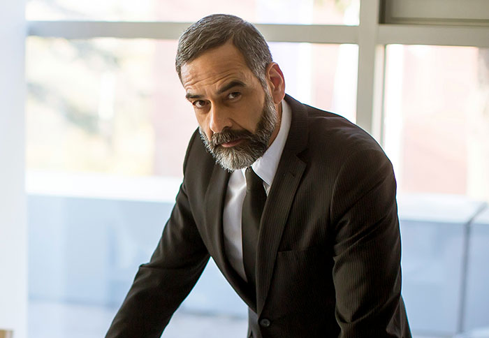 Middle-aged man in a black suit and tie looking confidently at the camera, embodying tall and loud boss presence.