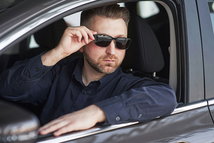 Man wearing sunglasses sitting in a car, projecting confidence and a tall, loud presence to entitled bosses.