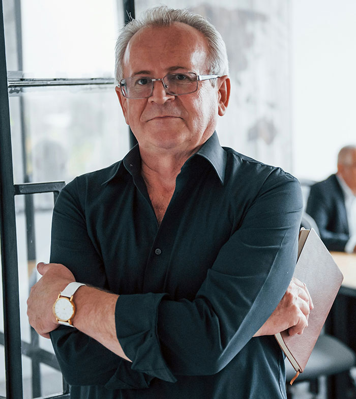 Older man with glasses in a black shirt holding a notebook, showing confidence in an office setting with entitled bosses.
