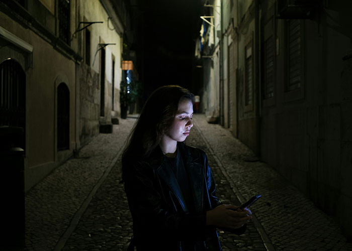 Woman standing alone on a dark street at night, using her phone cautiously, illustrating safety tips for women.