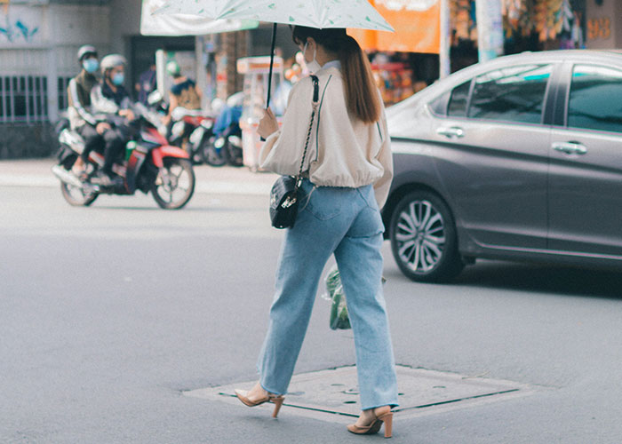 Woman crossing street with umbrella, wearing mask and heels, demonstrating safety tips women use in urban environments today