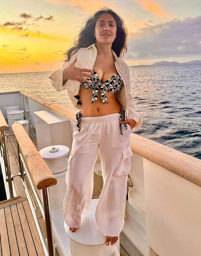 Salma Hayek models a black and white bikini top with white pants on a yacht at sunset for Sports Illustrated swimsuit cover.