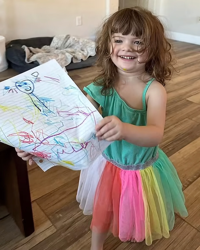 Toddler girl in colorful dress smiling and holding a drawing inside a home with wooden flooring and a dog bed. Toddler girl in colorful dress smiling and holding a drawing inside a home with wooden flooring and a dog bed.