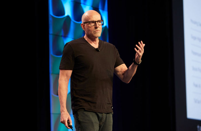 Scott Galloway presenting on stage, wearing glasses and black t-shirt, discussing money rules and net worth strategies.
