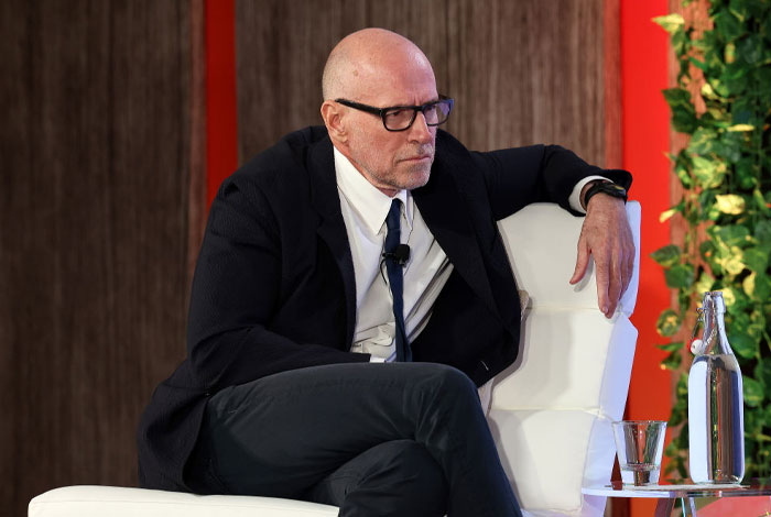 Scott Galloway in a suit and glasses seated on a white chair, engaged in a discussion about money rules and net worth.