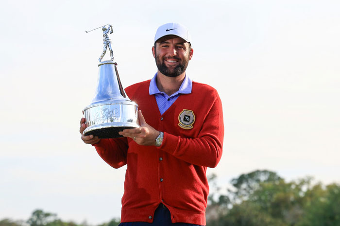 Scottie Scheffler holding golf trophy, wearing red sweater and white cap, showcasing financial power in golf.