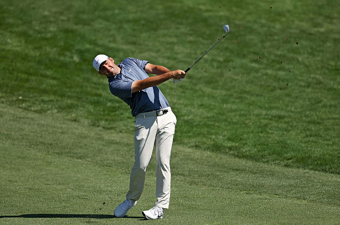 Golfer Scottie Scheffler in mid-swing on a green golf course showcasing golf&rsquo;s financial power and success.