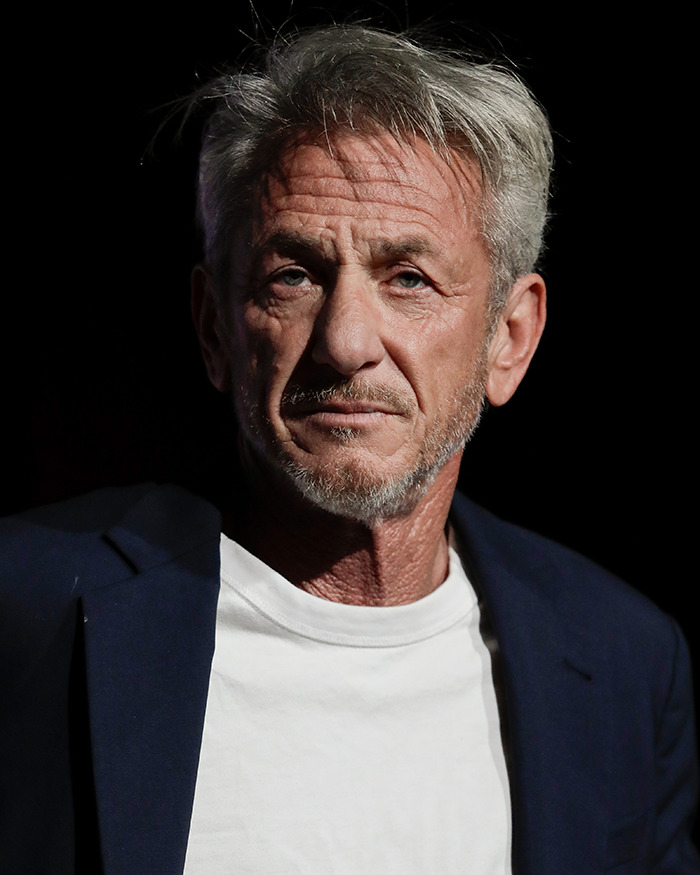 Sean Penn with a rough appearance, wearing a white shirt and dark blazer, photographed against a black background.