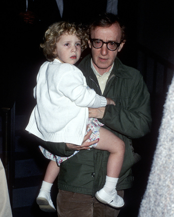 Woody Allen wearing glasses and a green jacket holding a young child in a white sweater at night.