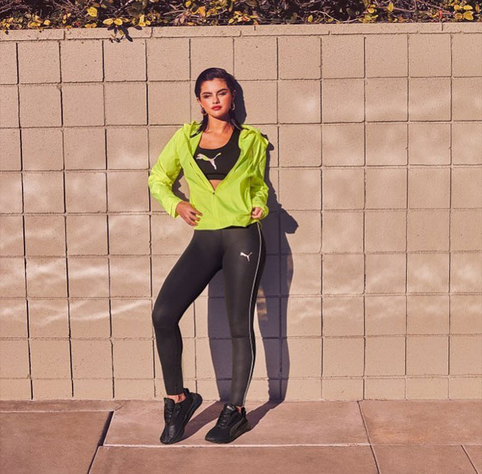 Selena Gomez in sporty outfit posing against a wall, highlighting her net worth from makeup and music earnings in 2025