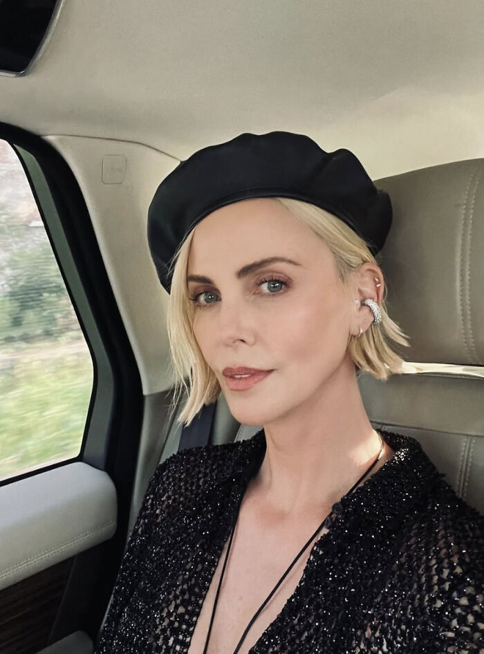 Charlize Theron wearing a black beret and sparkly top, sitting in a car and posing for a selfie.
