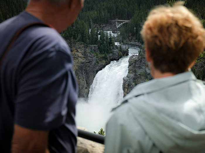 Two people observing a large waterfall in a forested area, showcasing natural beauty and outdoor scenery.