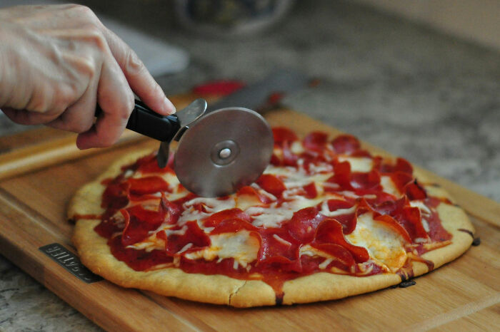 Hand using a pizza cutter on a pepperoni pizza, illustrating moments of people being surprisingly stupid.