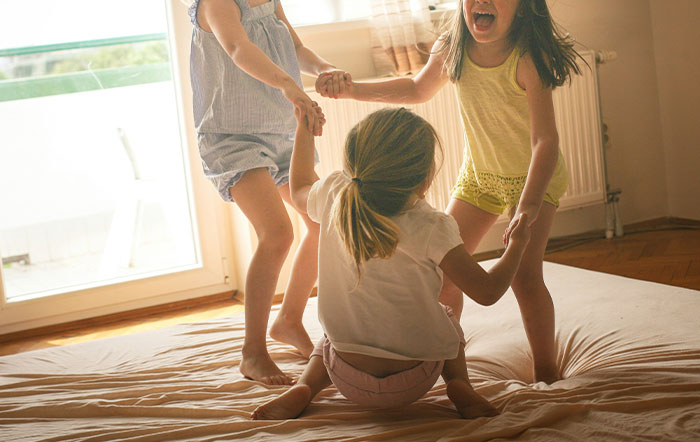 Three children holding hands and playing indoors on a bed, illustrating moments of unexpected behavior and foolishness.