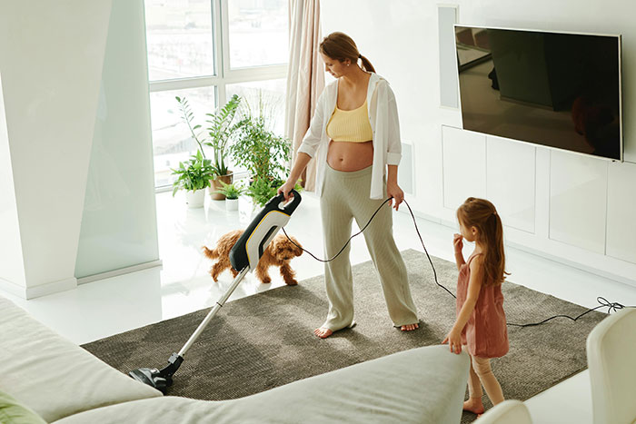 Woman vacuuming carpet in living room with child and dog nearby, portraying cleaning after sick husband vomits on floor. Woman vacuuming carpet in living room with child and dog nearby, portraying cleaning after sick husband vomits on floor.