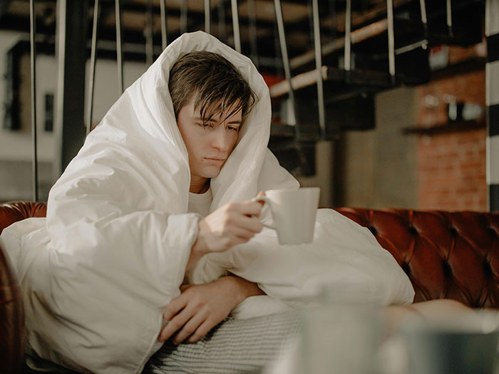 Sick husband wrapped in blanket looking unwell, holding a cup, refusing to use trash can to throw up at home.