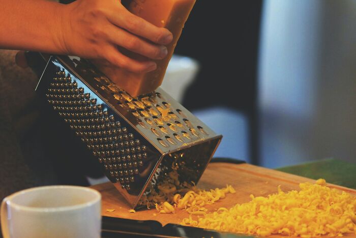 Person grating cheese with a box grater on a wooden board, highlighting foods that go bad more quickly.