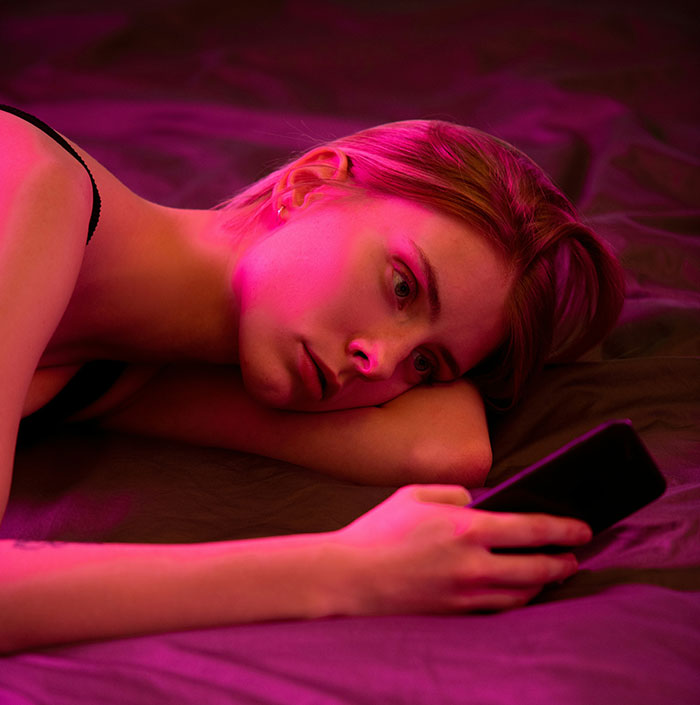 Young woman lying on bed under pink light, looking sad and distant while holding phone, reflecting signs of being cheated on.