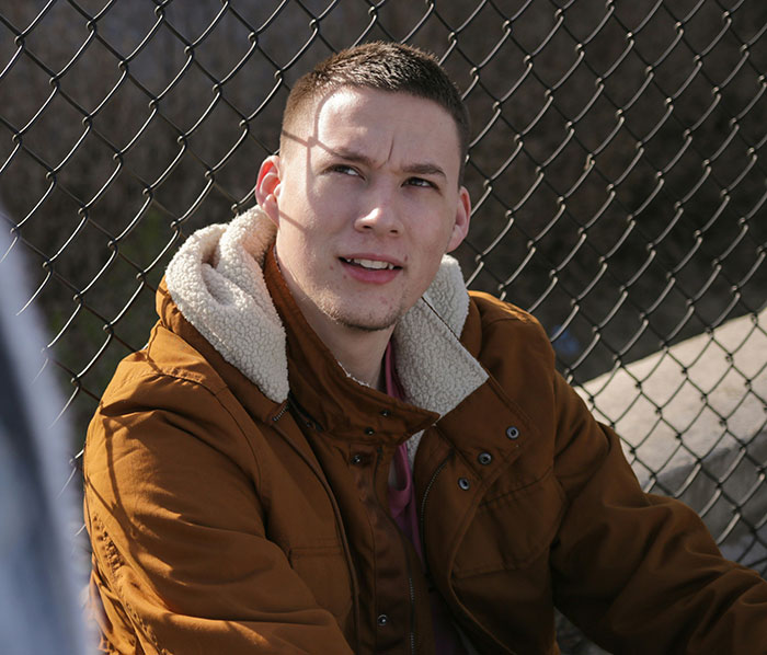 Young man in a brown jacket sitting by a chain-link fence, appearing thoughtful about signs of being cheated on.