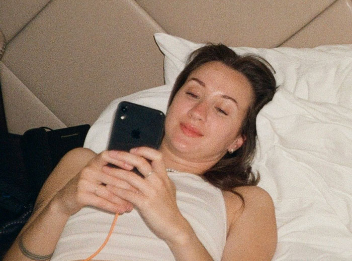 Young woman lying in bed, looking at her phone with a slight smile, showing subtle signs of pulling away in a relationship.
