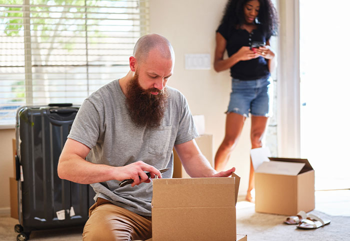 Man unpacking a box in a living room while a woman stands in the background using a phone, highlighting irresponsible parenting issues.