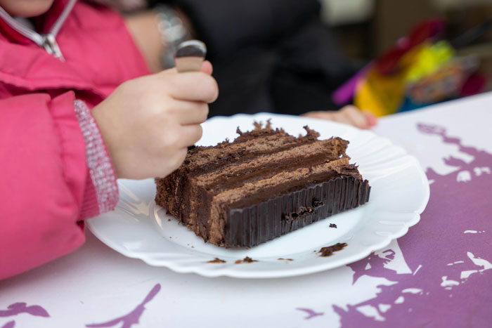 Child in pink jacket holding fork, about to eat a piece of chocolate cake at a birthday gathering with visiting relatives. Child in pink jacket holding fork, about to eat a piece of chocolate cake at a birthday gathering with visiting relatives.